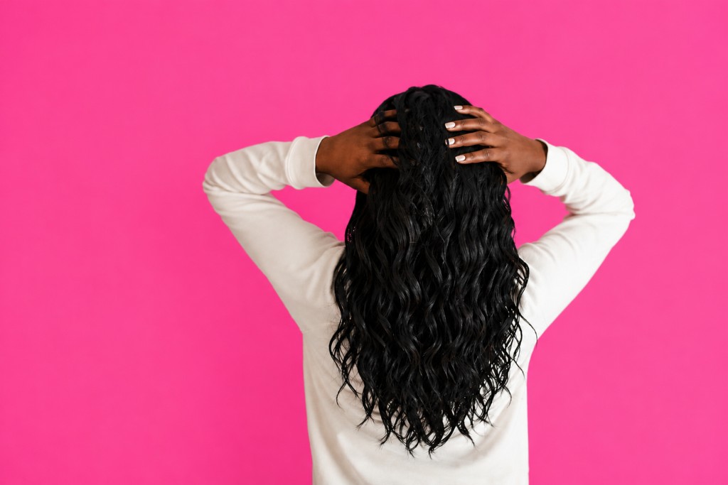 Back view of a person with long wavy black hair, hands in hair, cream top on a vibrant pink studio backdrop